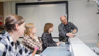 Kolme lukio-opiskelijaa seuraa, kun opettaja näyttää heille pöydällä olevan teknisen laitteen toimintaa.