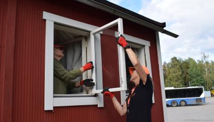 Punaisen talon ikkuna on auki ja kaksi henkilöä korjaavat sitä.