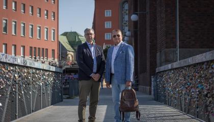 Jari Ahola ja Harri Airaksinen seisovat vierekkäin kevyenliikenteen sillalla, jonka kaiteisiin on kiinnitetty kymmeniä rakkauslukkoja, ja taustalla näkyy tiilenpunaisia korkeita rakennuksia..