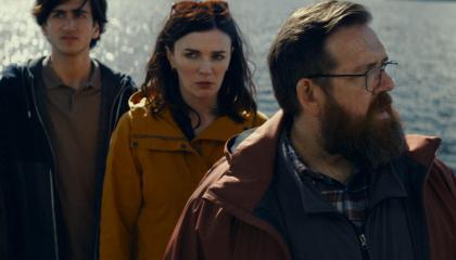 Vakavat näyttelijät seisovat laiturilla järvimaisema taustanaan.