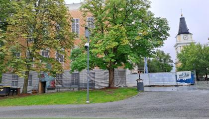 Suurten puiden takaa pilkottaa keltainen rakennus. Taustalla kohoaa kirkon tapuli.