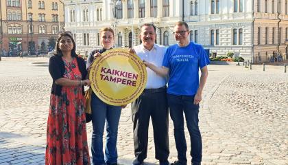 Neljä henkilöä, jotka esiintyvät Tampereella järjestettävässä rauhanseminaarissa. Yhdellä on käsissään iso pyöreä kyltti, jossa tulee Kaikkien Tampere.