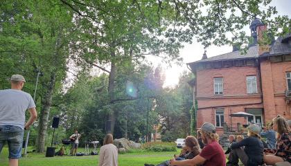 Muusikko esiintyy puistossa kesällä. Kuuntelijat istuvat nurmikolla.