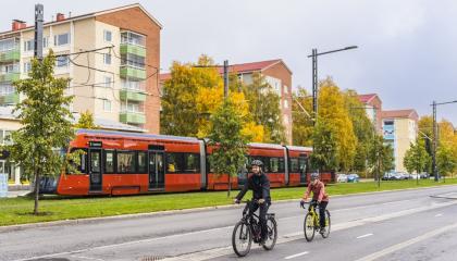 Kaksi henkilöä pyöräilemässä punaisen ratikan ajaessa taustalla.