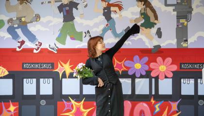 Muraalin tehnyt henkilö seisoo teoksensa edessä. Teoksessa on punainen raitiovaunu, jonka katolla juoksee nuoria. 