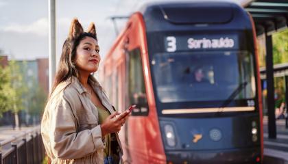 Nainen ja Ratikka ratikkapysäkillä. Naisella kännykkä kädessään ja hassut korvat päässä.