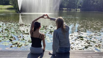 Kaksi tyttöä istuu Sorsapuiston lammen rannalla ja muodostaa sormilla yhdessä sydämen. 