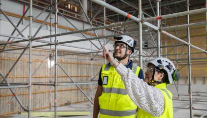 Kaksi henkilöä katselee valtuustosalin seinää. Toinen osoittaa kädellään katsesuuntaan. Kuvan tausta on täynnä rakennustelineitä.