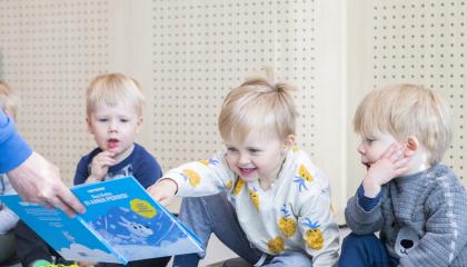 Kolme poikaa istuu lattialla ja tutkii satukirjaa, jota aikuinen näyttää heille.