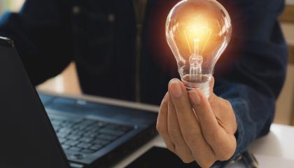 Ihminen istuu tietokoneen ääressä ja pitää kädessään hehkulamppua, jossa on valo.