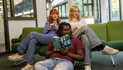 Nuoria kirjastossa. Etualalla poika lukemassa kirjaa. Takana olevalla sohvalla kaksi tyttöä pelaamassa pelikonsolilla.