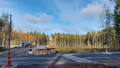Katu, jonka takana metsän ympäröimä aukio. Keskellä katutyöpuomeja ja niiden takana kuorma-auto.