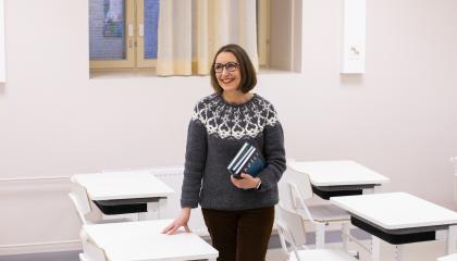 Opettaja seisoo luokassa pulpettien keskellä kirjoja kädessään.