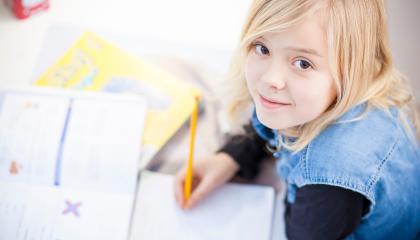 Vaaleahiuksinen lapsi on tekemässä läksyjä koulupöydän ääressä. Hän on nostanut katseensa kohti katsojaa.