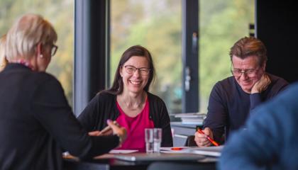 Hymyileviä ihmisiä istuu pöydän ääressä. Pöydällä tietokone ja papereita.