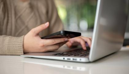 Henkilön kädet, joista toinen on kannettavan tietokoneen näppäimistöllä ja toinen pitelee älypuhelinta.