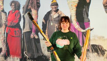 Nainen pitelee käsissään kahta isoa puista soitinta, taustalla aboriginaalitaiteilijoita esittävä kuva.