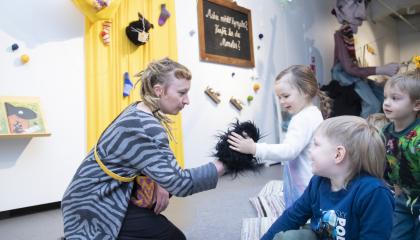 Ohjaaja näyttää lapsille pörröistä mustaa leikkilemmikkiä. Yksi heistä on kohottautunut polvilleen silittämään sitä, ja muut katsovat.