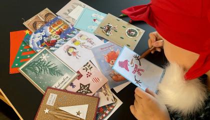 Lapsi tonttulakissa piirtämässä joulukorttia. Pöydällä kortteja.