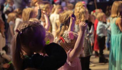 Juhla-asuinen lapsi katselee edessään olevia muita lapsia, jotka tanssivat. 