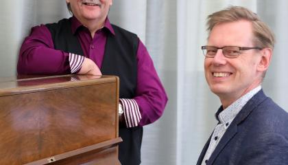 Sinitakkinen mies istuu pianon ääressä, toinen violettipaitainen mies nojaa pianoon taaempana. Molemmat hymyilevät.