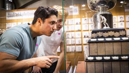 Tummatukkainen mies osoittaa vitriinissä olevaa suurta jääkiekkopokaalia, vaaleahiuksinen mies taustalla.