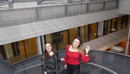 Kaksi henkilöä hymyilee iloisina metallisissa portaissa. Toinen vilkuttaa.