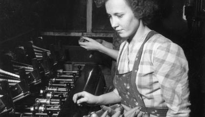 Mustavalkoinen kuva Finlaysonin puuvillatehtaan kutojasta 1930-luvulla. Ruutupaitainen nainen, jolla on käsissään laatikollinen lankapuolia.