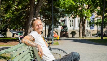 Mies istuu rennosti puistonpenkillä ja hymyilee, taustalla isoja puita.