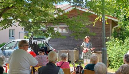 Nainen soittaa ukulelea kesäisessä puutarhassa ja tuoleilla istuvat istuvat ikäihmiset laulavat mukana.