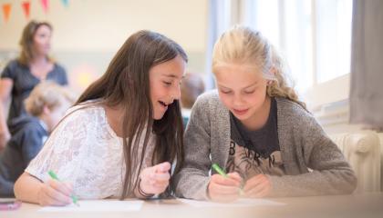 Kaksi nuorta naisoletettua istuu pöydän ääressä kynät kädessä.