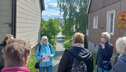 Sinitakkinen nainen opastaa ulkovaatteisiin pukeutuneita ihmisiä Viinikan puutalojen välissä kulttuuriraitilla.