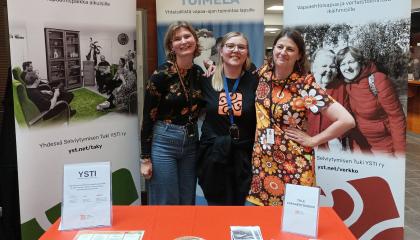 Kolme naista seisoo hymyillen esittelytiskin takana messutilaisuudessa.