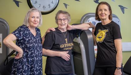Kolme seniori-ikäistä hymyilevää rouvaa sseisoo vierekkäin kuntosalilla.