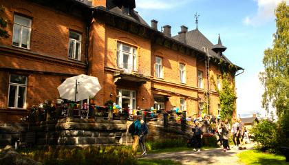 Punatiilinen kartanorakennus kesäisenä päivänä, terassilla ja edustalla ihmisiä.