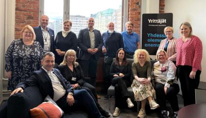 Kolmentoista hengen porukka ryhmäkuvassa. Osa istuu sohvilla, loput seisovat. Kaikki hymyilevät. 