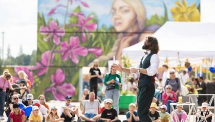 Mies esiintyy joukolle ihmisiä kesäisenä päivänä. Taustalla värikäs seinämalaus.