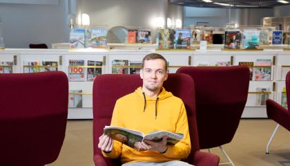 Henkilö istuu nojatuolissa pääkirjasto Metson lehtisalissa ja pitää käsissään avointa lehteä. Taustalla on salin lehtikalusteita. 