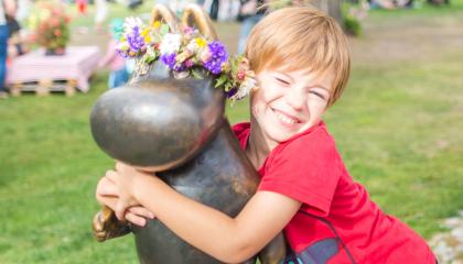 Punapaitainen poika halaa Muumipeikko-patsasta, jolla on kukkaseppele päässä. Taustalla ihmisiä kesäisessä puistossa.