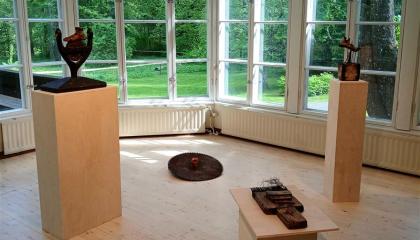 Eri kokoisia tummia veistoksia näyttelysalissa. Taustalla suuret ruutuikkunat, joista aukeaa vehreä kesäinen maisema.