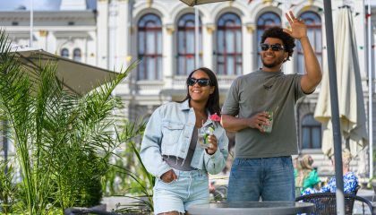 Kaksi aurinkoisesti hymyilevää ihmistä seisoo vehreällä Keskustorin kesän terassialueella, ja kuvan taustalla näkyy Raatihuone.