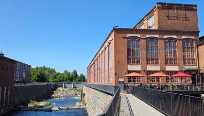 Vapriikin punatiilinen rakennus ulkoa kuvattuna. Edustalla ravintolan ulkoterassi, jossa varjoja ja ruokailijoita. Rakennuksen reunassa alapuolella virtaa koski.