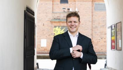 Henkilö seisoo kädet edessään ulkona valkoisten seinien välissä takanaan tiiliseinää.