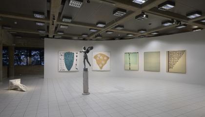 Vaaleaseinäinen näyttelytila, jossa keskellä harmaa patsas. Taustalla näkyy suuria haaleasävyisiä maalauksia.