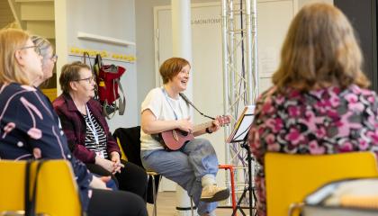 Nainen laulaa ja soittaa ukuleleä kolmen muun naisen kuunnellessa.