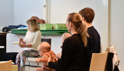 Kaksi henkilöä istuu tuolillaan vauva sylissä. Taustalla muskariopettaja.