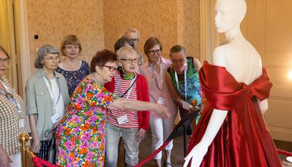 Naiset seisovat museonäyttelyssä ja katselevat punaista, olkaimetonta silkkisatiinista valmistettua iltapukua.