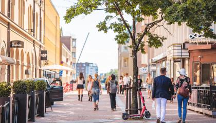 Ihmisiä kävelee kesäaikaan kadulla. Etualalla puu, johon nojaa sähköpotkulauta.