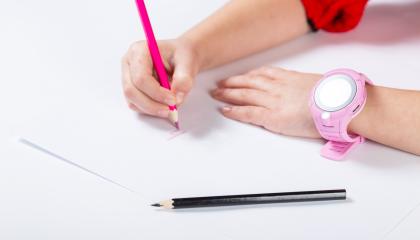 Nuori käsi kirjoittaa punaisella värikynällä paperille. Toisessa kädessä on vaaleanpunainen älykello