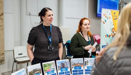 Kaksi Tampereen kaupungin asiantuntijaa keskustelee asukasillassa ihmisten kanssa. Pöydällä on useita esitetelineitä.